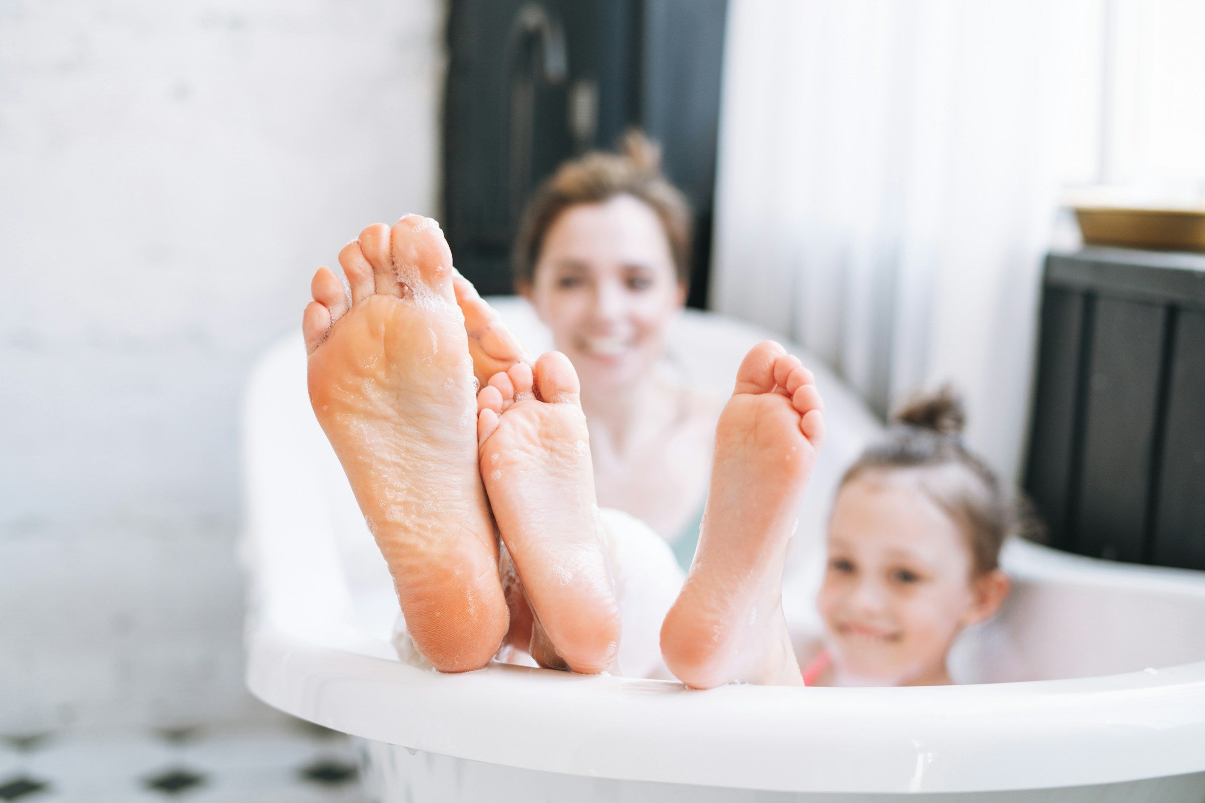 Two pairs of feet in a bathtub with a blurred background of a woman and child.